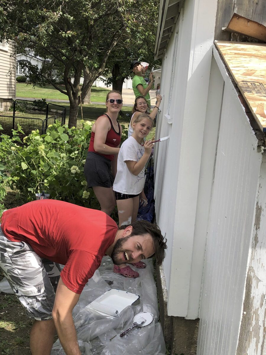 Five people painting the side of a house outdoors, surrounded by greenery. They are smiling and holding paintbrushes.