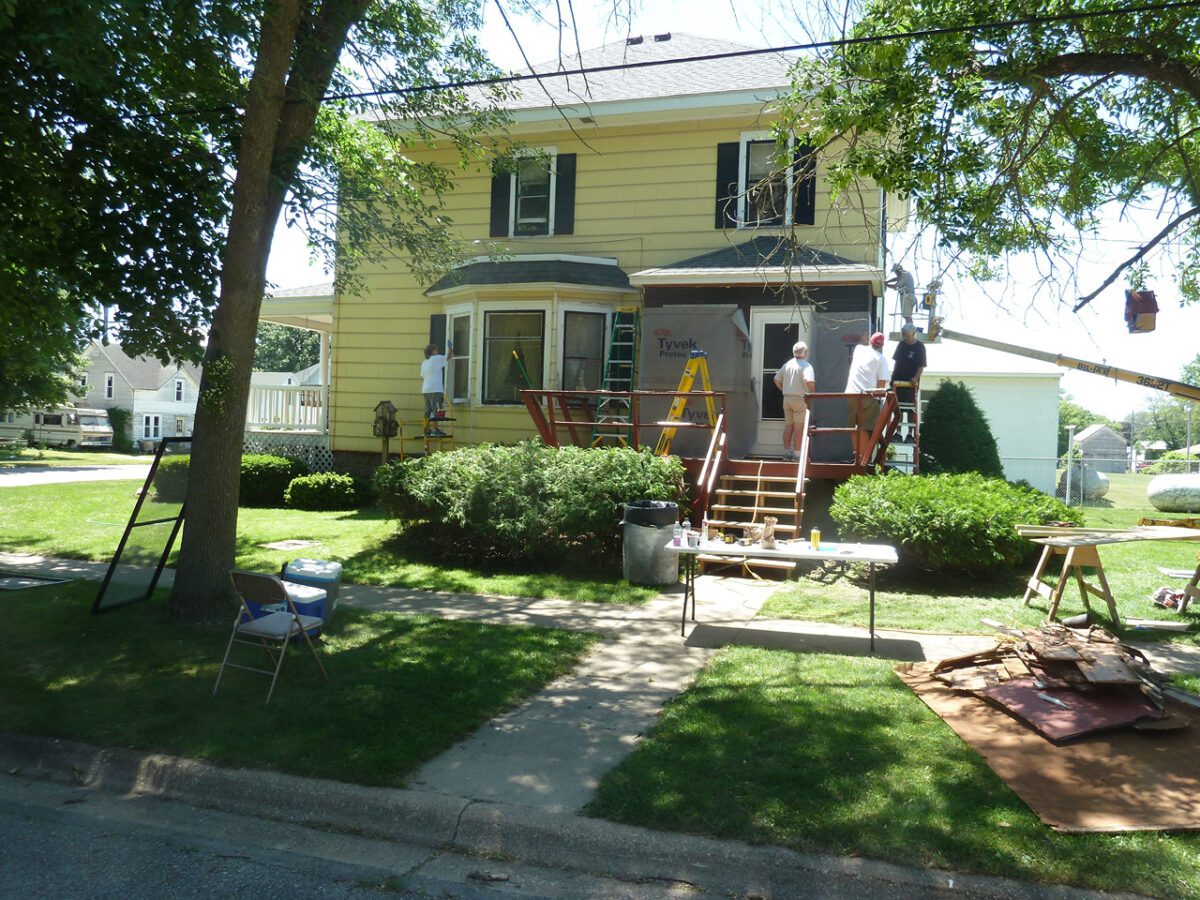 People working on the exterior of a yellow two-story house with a front porch, surrounded by trees and grass.