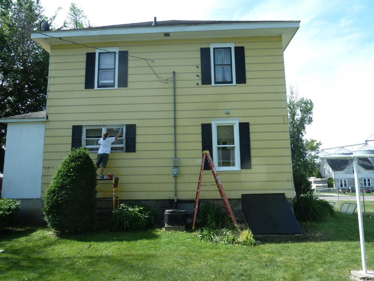 Person stands on scaffolding painting window frame on yellow two-story house with black shutters. Red ladder is nearby on grass.