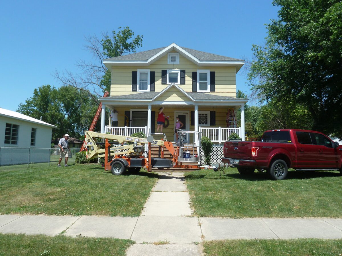 A yellow house with white trim, a front porch, ladder, and construction equipment on the lawn. A red truck is parked in the driveway, and two workers are near the house.