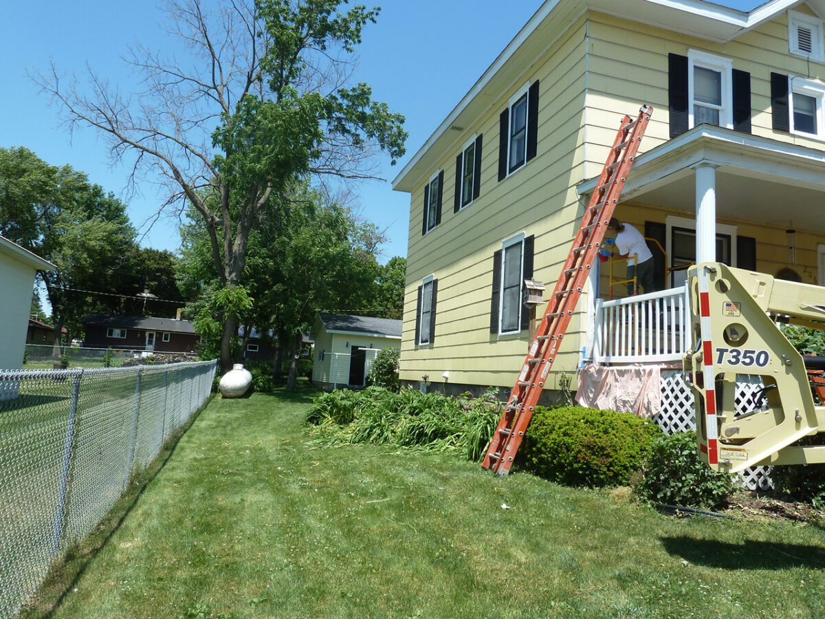 A yellow house with a ladder leaning against it. A lift vehicle is parked on the lawn, and a person is on the front porch. A chain-link fence runs along the side.