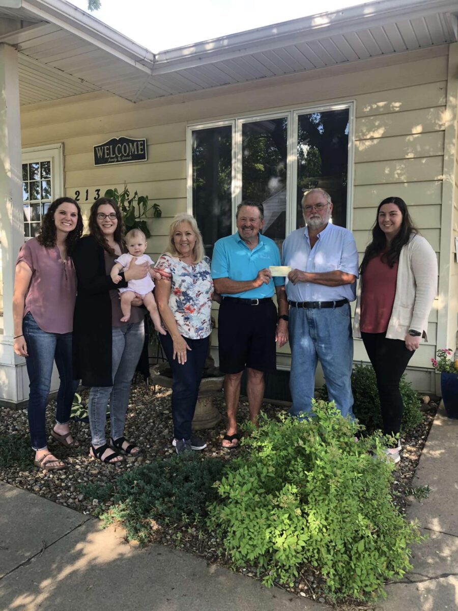 Group of people standing together, including women, a man, and one holding a baby, in front of a house with a 