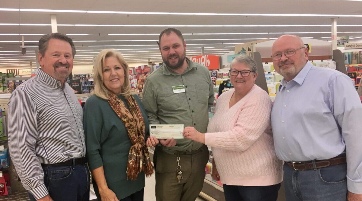 Five people stand in a store aisle; one person hands a check to another.