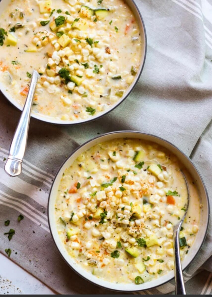 Two bowls of creamy vegetable soup with corn, zucchini, and herbs on a striped cloth, each with a spoon.